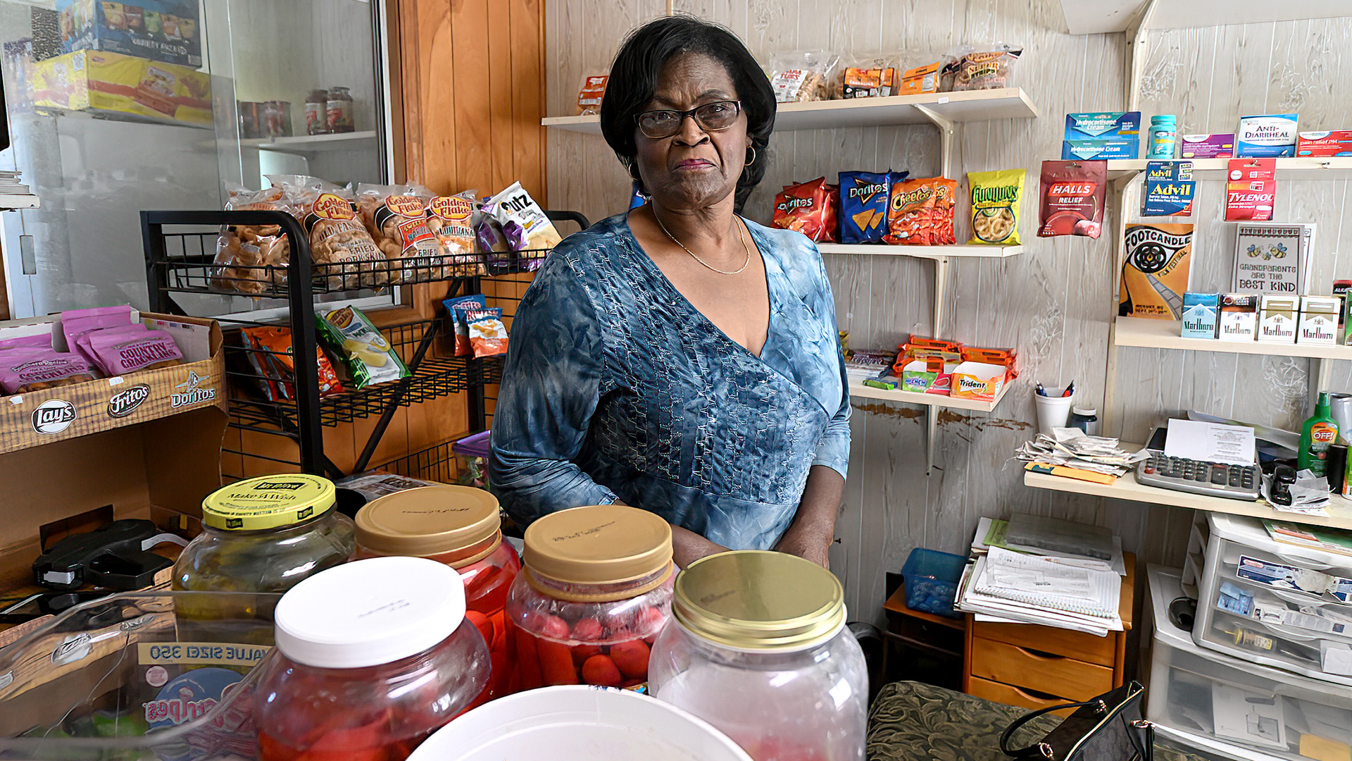 Dorothy Oliver poses for a portrait while standing behind a counter with multiple jars of food and in front of racks and wall-mounted shelves stocked with food, medications and cigarettes, with wood and plastic filing storage bins on the floor of a room with wood-paneled walls and an interior window.