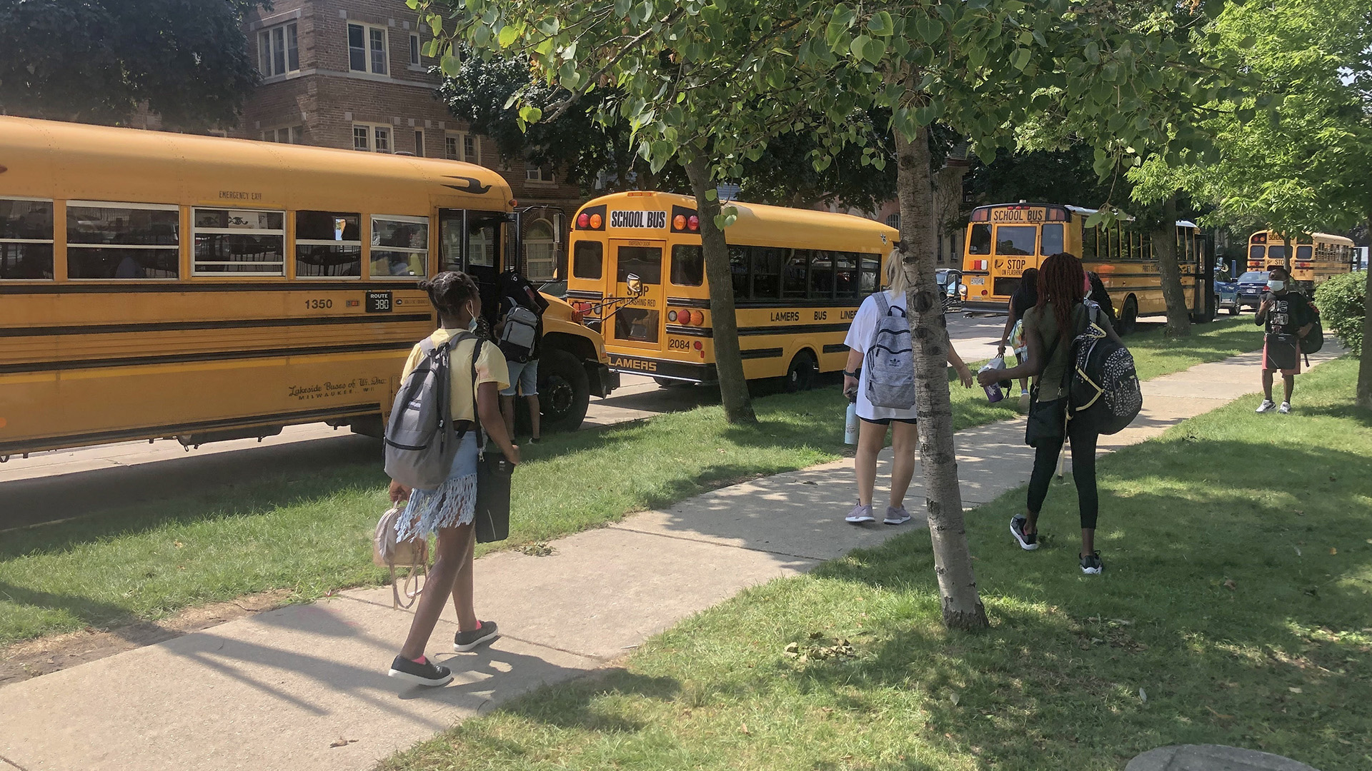Multiple people stand on a sidewalk and lawn in the sunlight and in the shade of trees, in front of a row of school buses parked in front of a brick building.