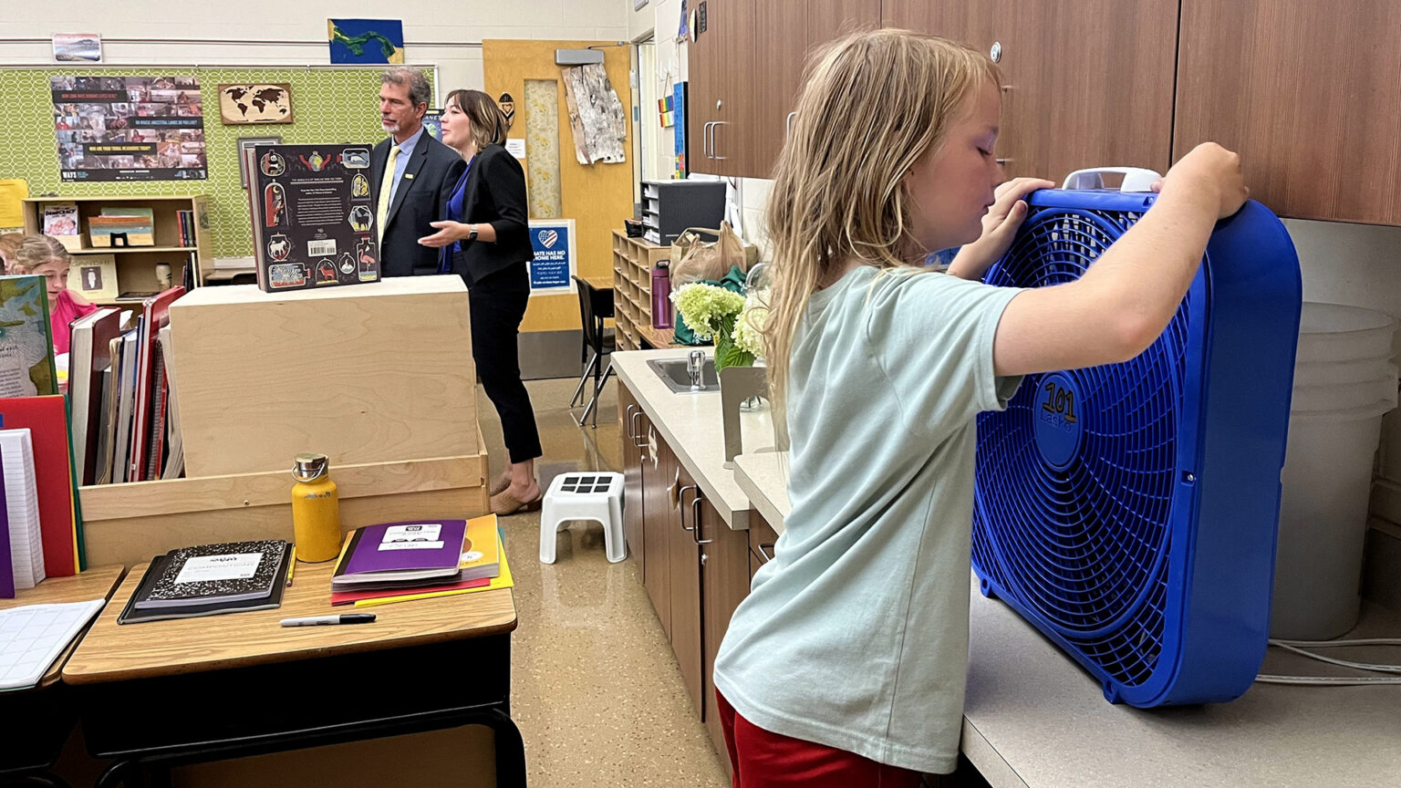 A child stands and turns on a box fan placed on a counter in classroom with cabinets, desks and posters attached to walls, with two adults standing in the background.