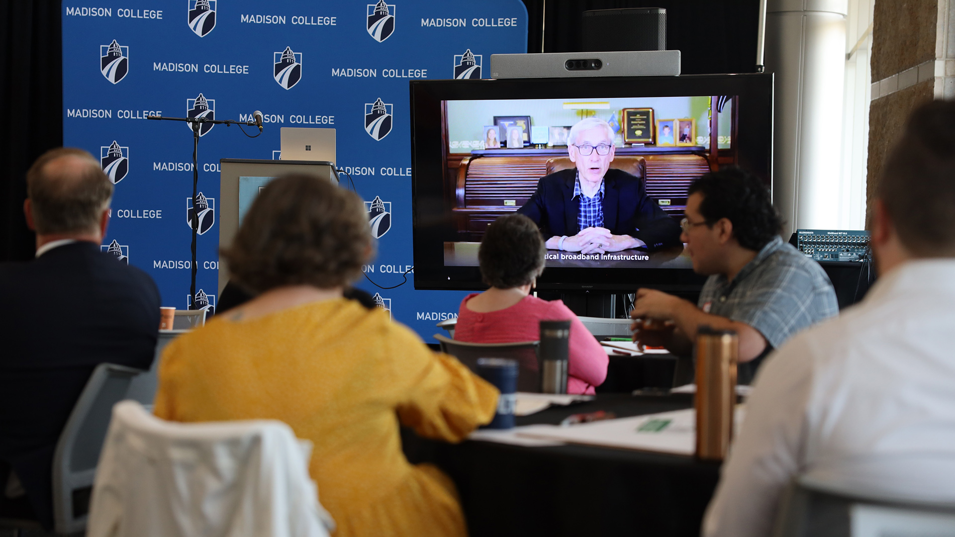 Multiple people seated in a room watch a video monitor showing Tony Evers speaking, in a room with a backdrop banner showing the words "Madison College" and its logo.