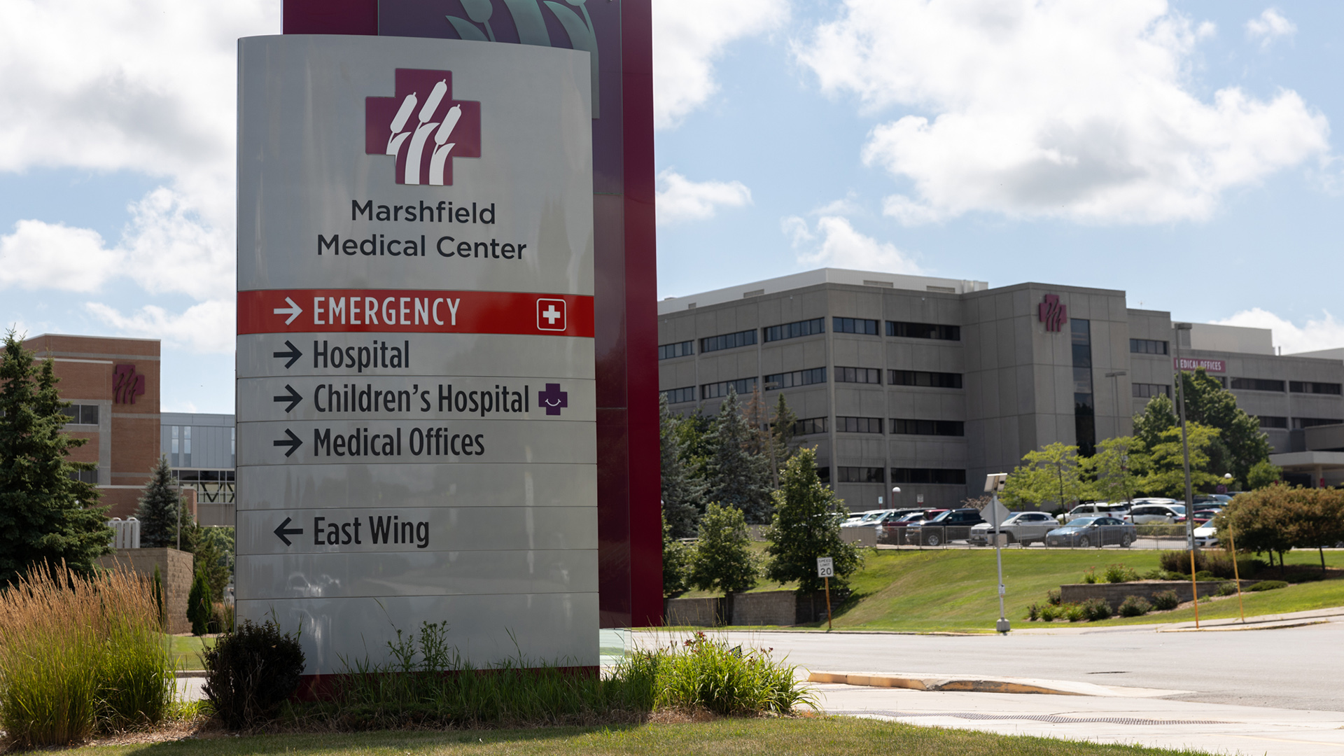 A direction sight for Marshfield Medical Center shows arrows pointing toward the locations for Emergency, Hospital, Children's Hospital, Medical Offices and East Wing, with a road, parking lot, trees and multi-story building in the background.