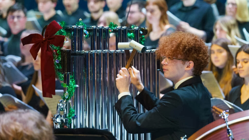 A young man plays tubular bells, or orchestral chimes, which are adorned with a red bow and green garland.