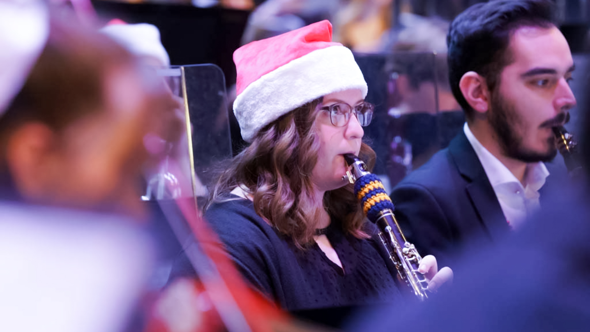 A young woman playing the clarinet in an orchestra wears a Santa hat.