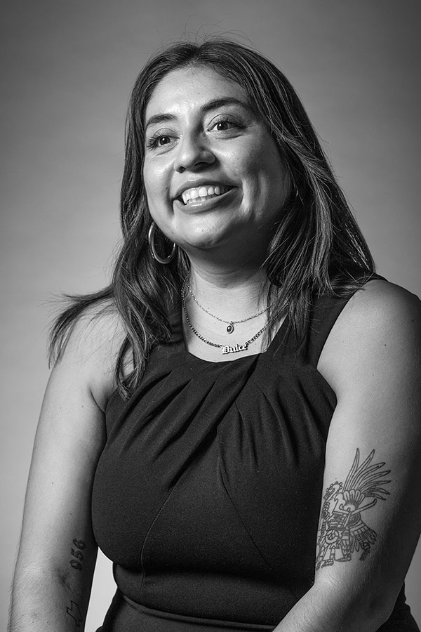 Woman smiling, wearing a black dress and necklace with the name "Dulce". She has a tattoo of the Aztec diety Huitzilopochtli on her arm.