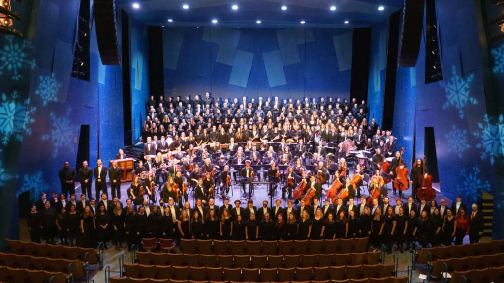 A college orchestra and choir poses on stage for a group portrait with blue lights and snowflakes decorating the backdrop.