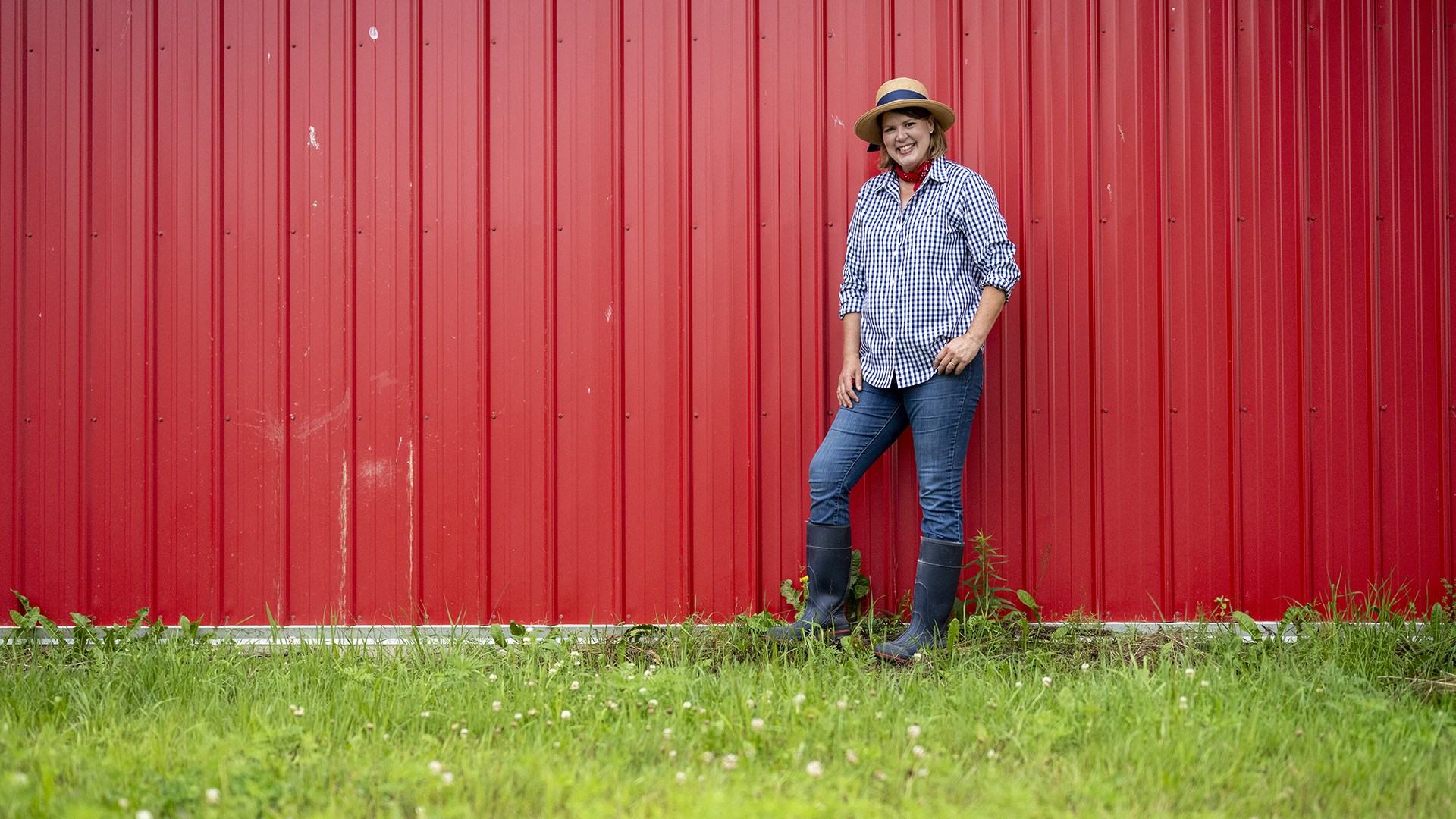"Around the Farm Table" host Inga Witscher wears a sun hat, button-up work shirt and rubber boots. She stands in front of a red barn on a summer day.