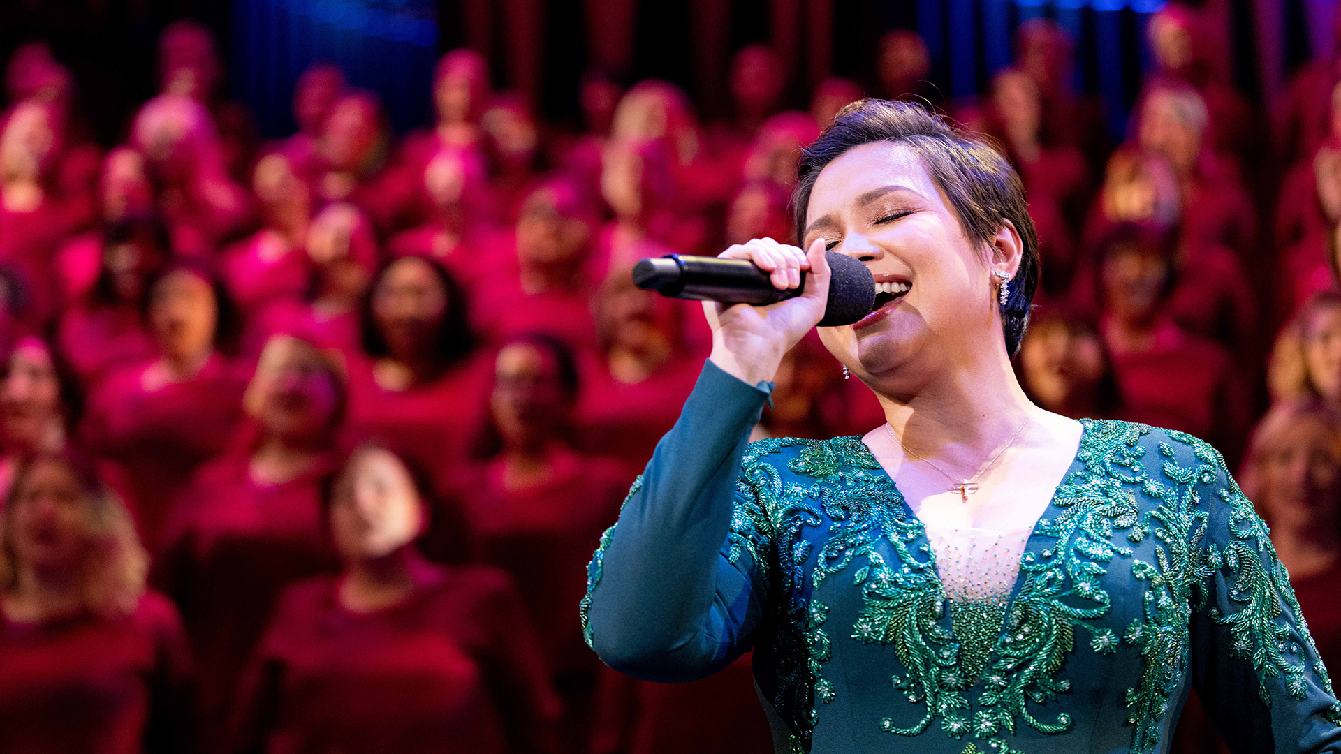 Singer Lea Salonga sings into a microphone while a choir dressed in red sings in the background.