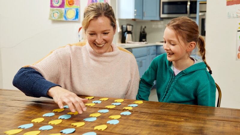 A mother and child arrange blue and yellow paper flowers in a pattern on a table,