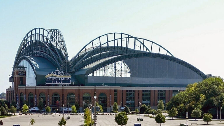 A retractable roof is in an open position on top of a baseball stadium with a sign over its home plate entrance reading American Family Field, with an empty parking lots and trees in the foreground.