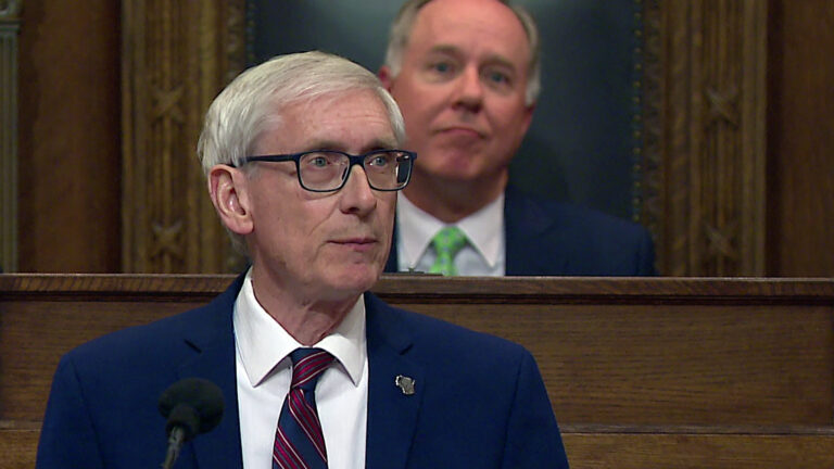 Tony Evers stands and speaks into a microphone with Robin Vos seated at a dais behind him.