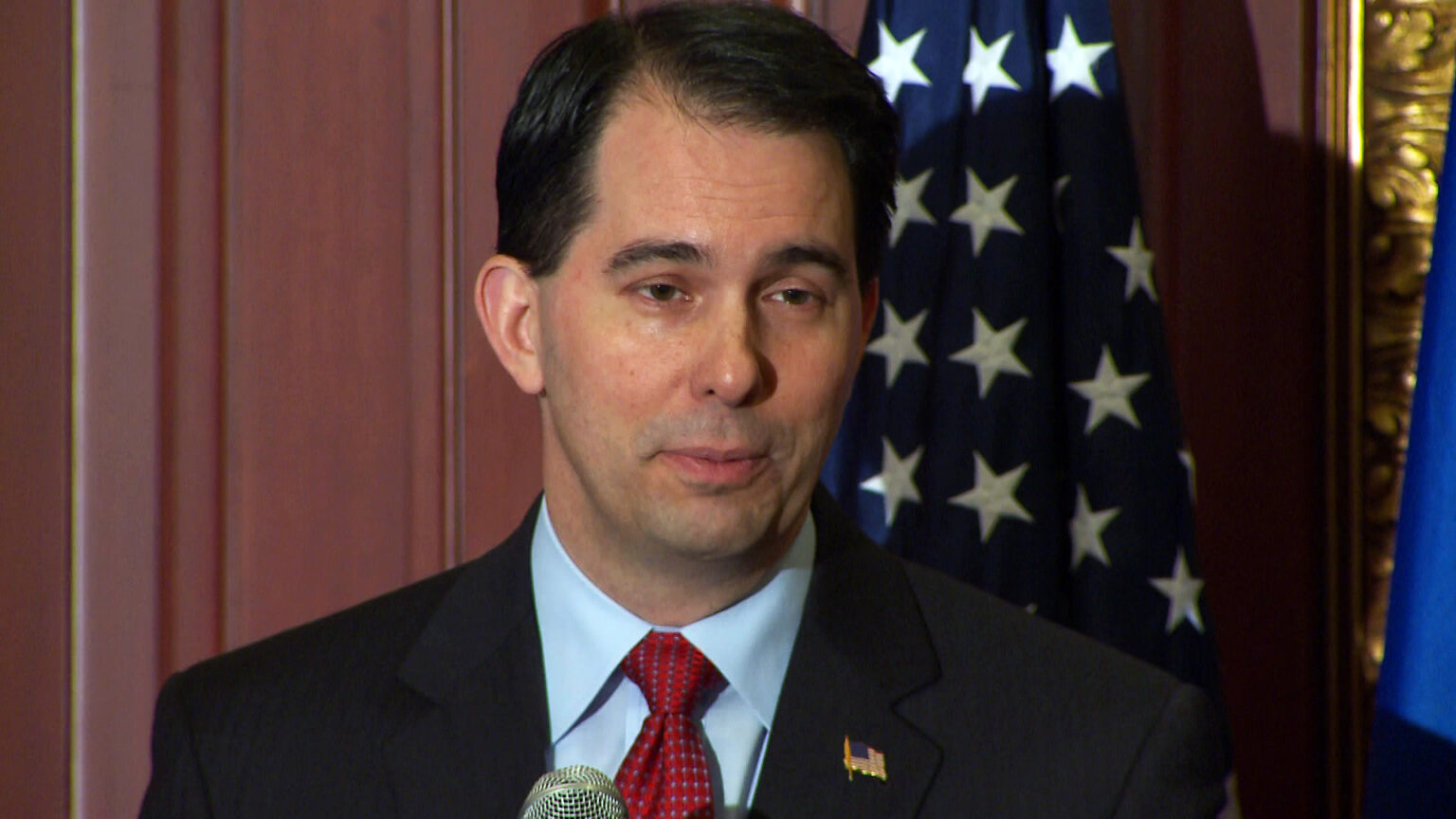 Scott Walker stands and speaks into a microphone while standing in front of a U.S. flag and wood wall paneling.