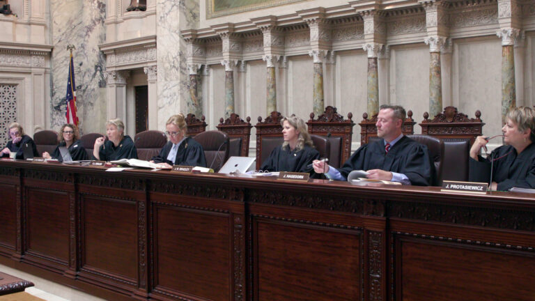 From left to right, Jill Karofsky, Rebecca Dallet, Ann Walsh Bradley Annette Ziegler, Rebecca Bradley, Brian Hagedorn and Janet Protasiewicz sit at a judicial dais in a row of high-backed leather chairs behind them, with another row of high-backed wood and leather chairs behind them, in a room with marble masonry and a U.S. flag.