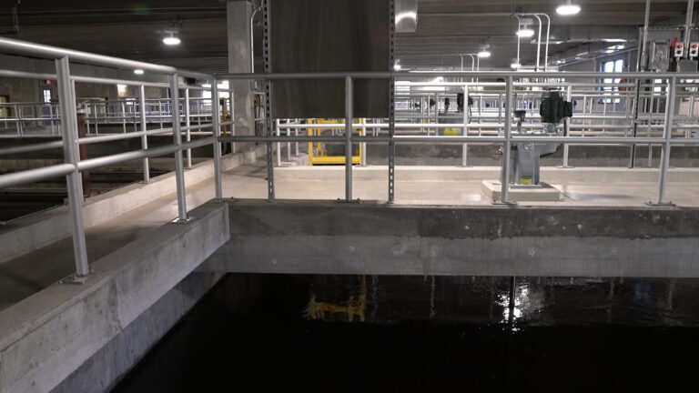 Overhead lights illuminate the interior concrete ceiling and walls of a wastewater treatment plant, with metal guard railings separating walkways from the surface of open tanks filled with water.