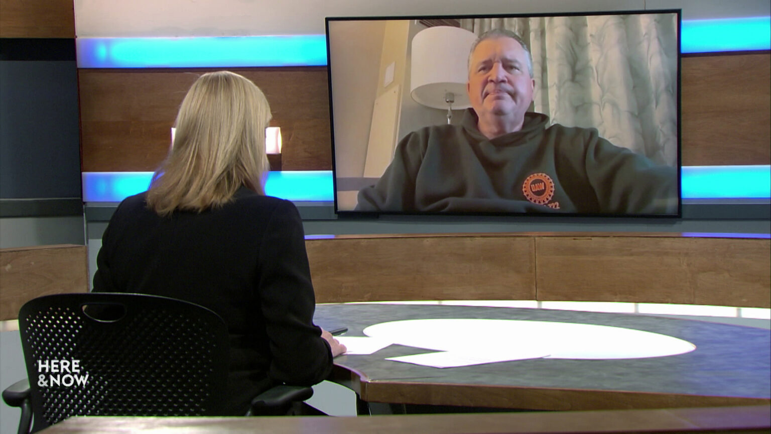 Frederica Freyberg sits at a desk on the Here & Now set and faces a video monitor showing an image of Steve Frisch.