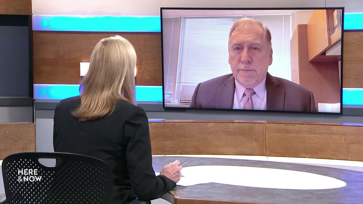 Frederica Freyberg sits at a desk on the Here & Now set and faces a video monitor showing an image of Don Millis.