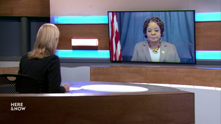 Frederica Freyberg sits at a desk on the Here & Now set and faces a video monitor showing an image of Gwen Moore.