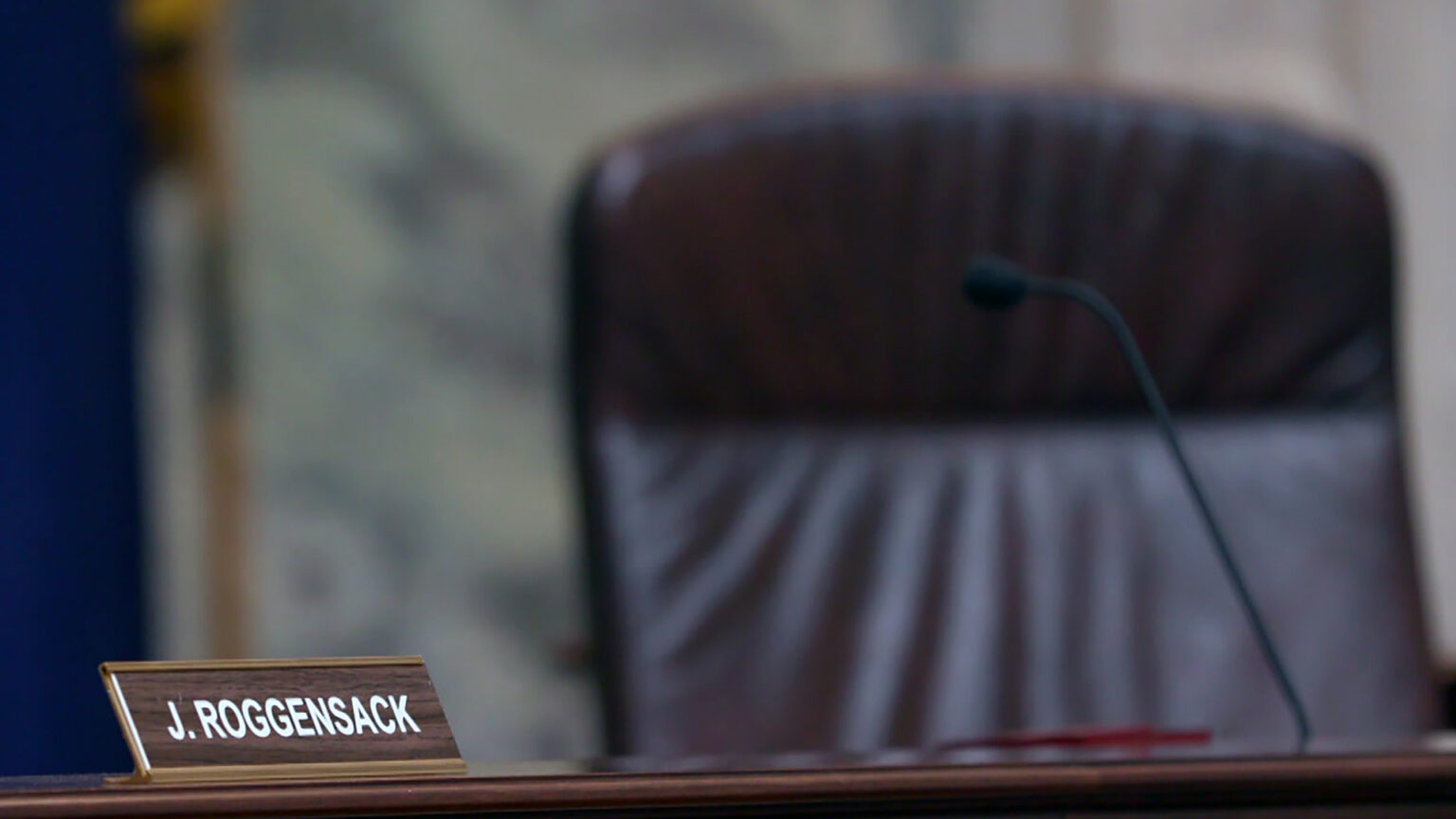 A nameplate reading J. Roggensack sits atop a wood judicial dais, with an out-of-focus microphone and high-backed leather chair in the background.