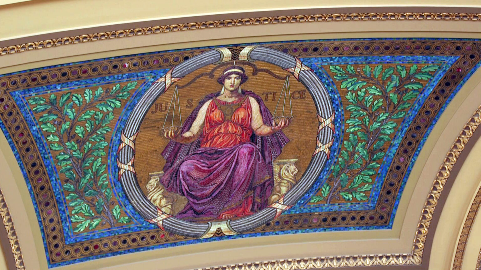 A mosaic depicting a Classical figure holding a scale and labeled Justice is seen on a curved interior wall of a rotunda inside a building.