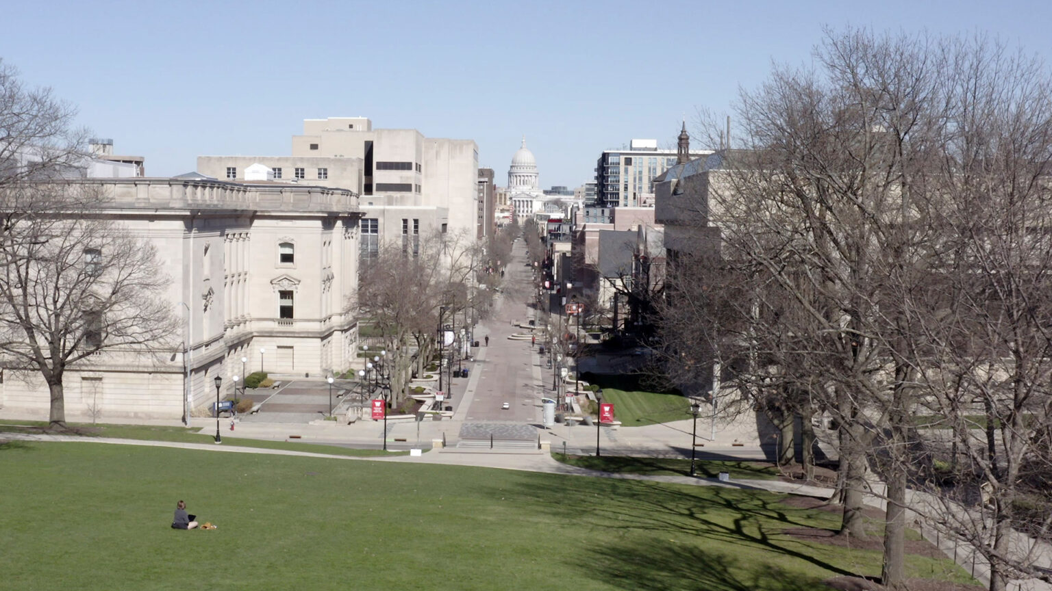 An aerial photo shows areas of grassy lawn at the bottom of a hill, with masonry and concrete buildings on either side of a street leading to the dome of the Wisconsin State Capitol, with leafless trees on both sides.