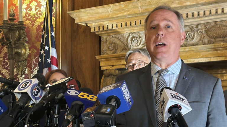 Robin Vos stands and speaks into multiple microphones equipped with flags of different media organizations, with other people standing behind him, in a room with a carved marble fireplace lintel, wood paneling, toile wallpaper, a brass wall sconce and a U.S. flag.