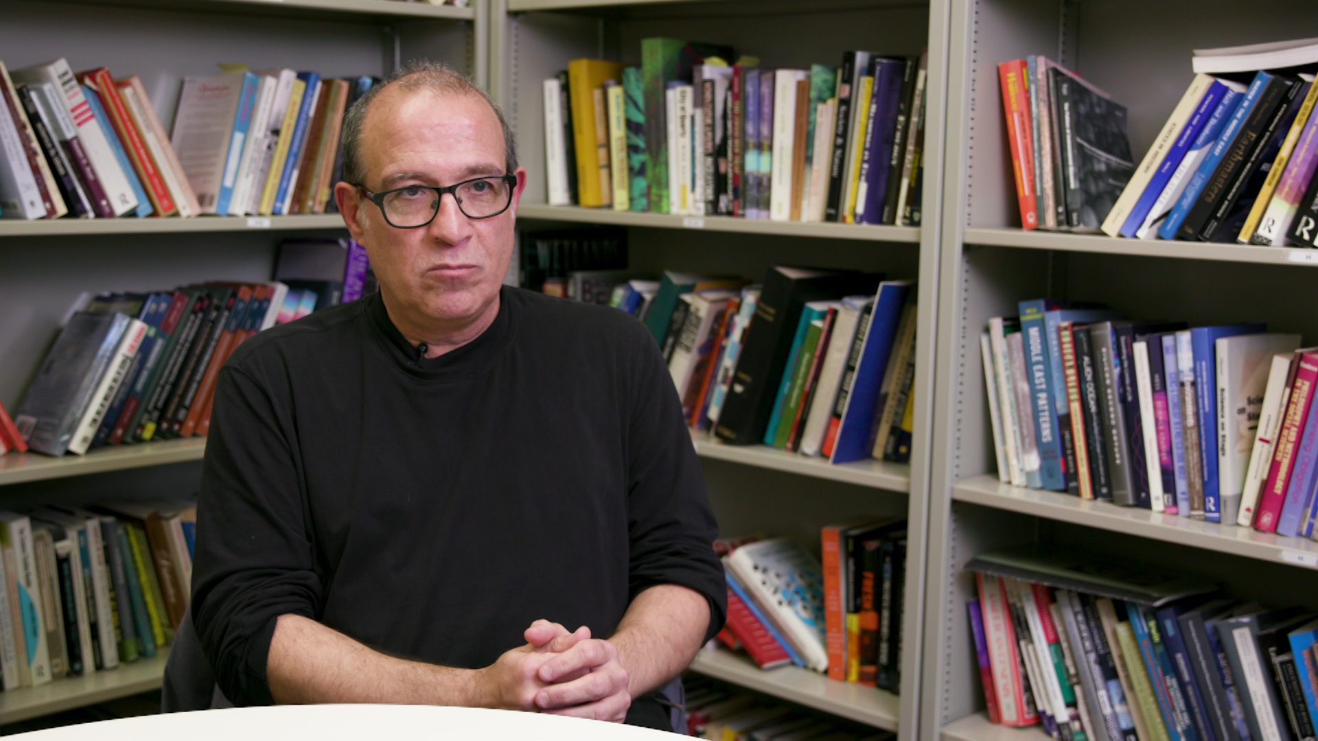 Samer Alatout sits at a table with his hands folded in his lap, with multiple metal bookshelves filled with books arranged at a right-angle behind him.