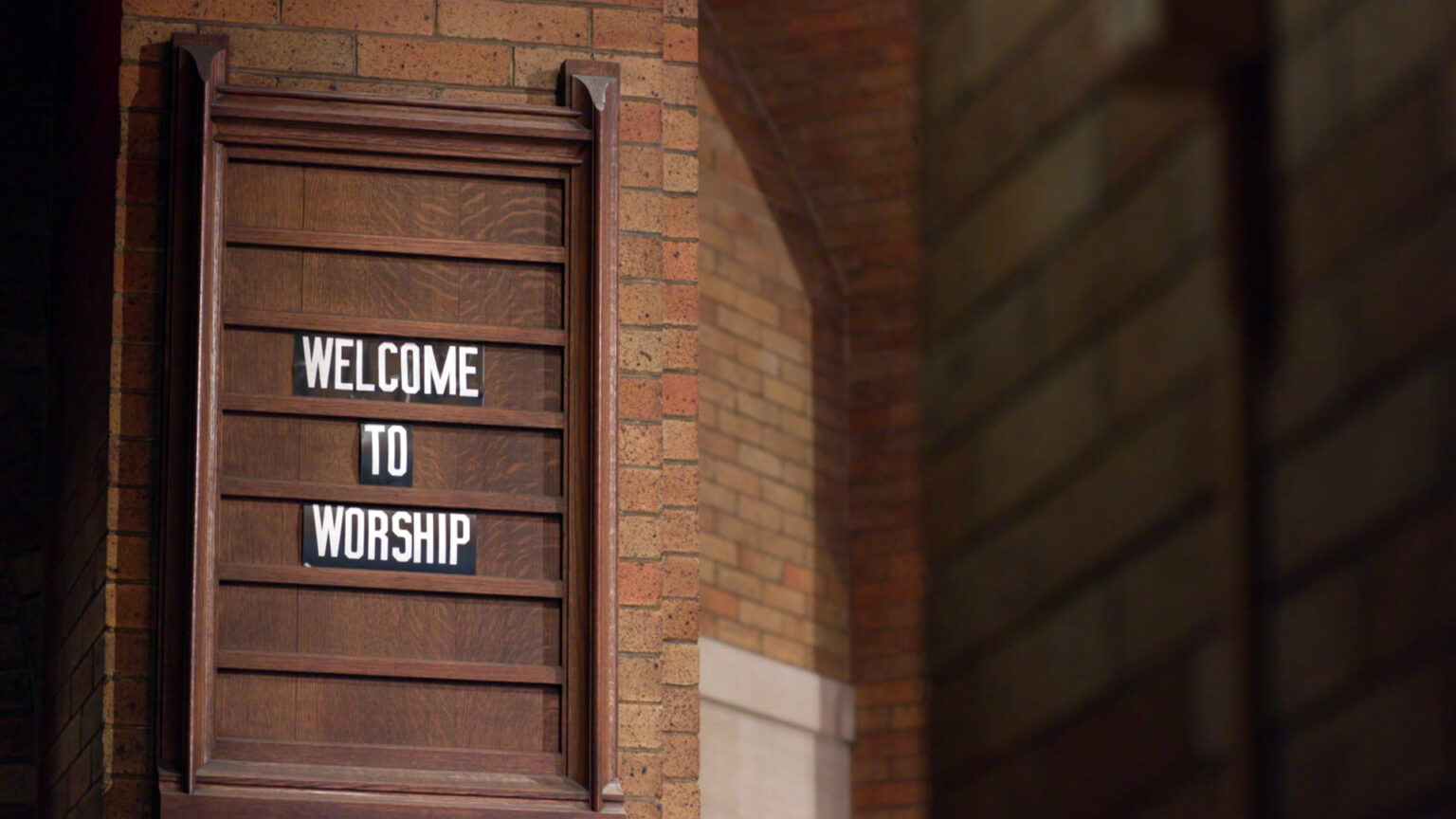 A wood sign with letters reading Welcome to Worship is mounted to a brick wall inside a building.