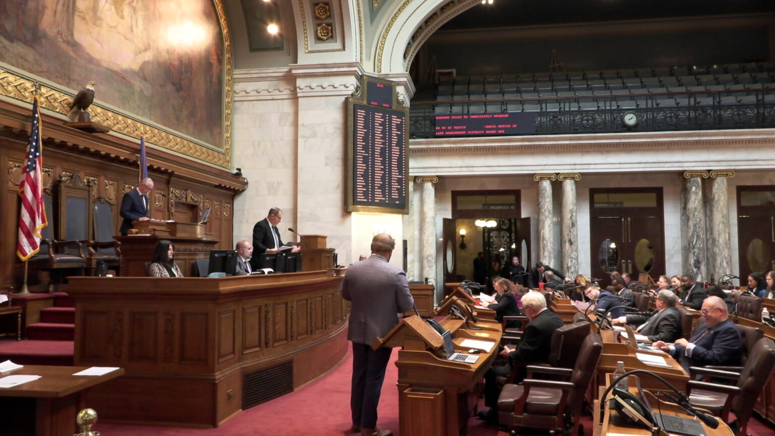 Lawmakers sit and stand in multiple rows of desks and a legislative dais in a room with marble pillars and masonry, a second-story seating gallery, the U.S. and Wisconsin flags, a taxidermy bald eagle, a large painting and a digital vote register.