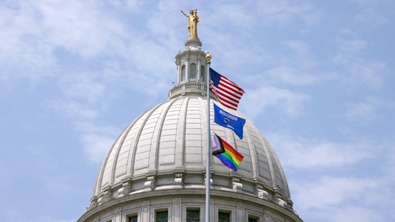 The U.S., Wisconsin, and Progress Pride flags wave in wind on a flagpole in front of the Wisconsin Capitol dome topped by a statue titled Wisconsin, with a partly cloudy sky in the background.