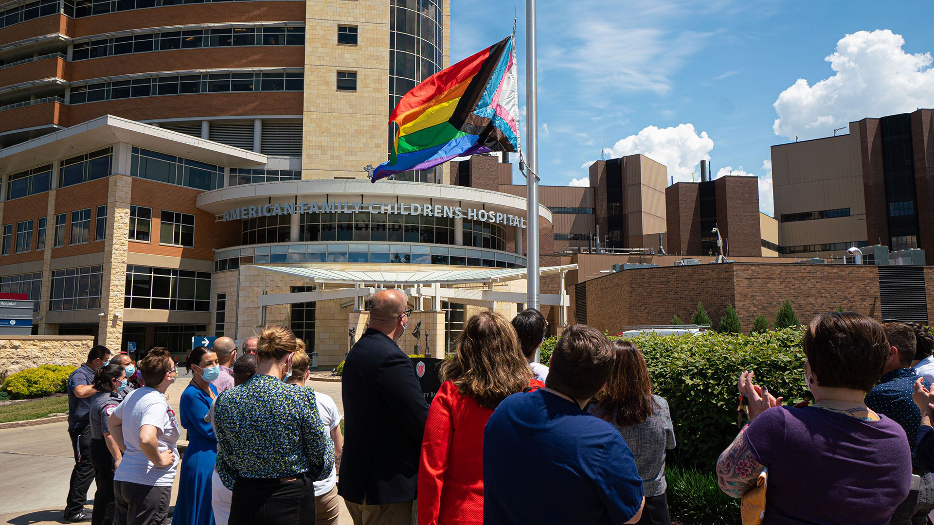 People watch and applaud as the Progress Pride flag is raised on a flagpole in front of a group of buildings, with a letter sign above one entrance reading "American Family Children's Hospital."