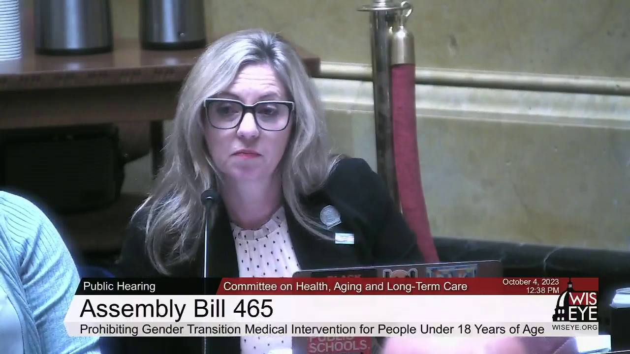 A video still image shows Robyn Vining sitting at a table and speaking into a microphone and facing an open laptop computer in a room with another person seated to the side, with a video graphic at bottom including the text "Assembly Bill 465" and "Prohibiting Gender Transition Medical Intervention for People Under 18 Years of Age."