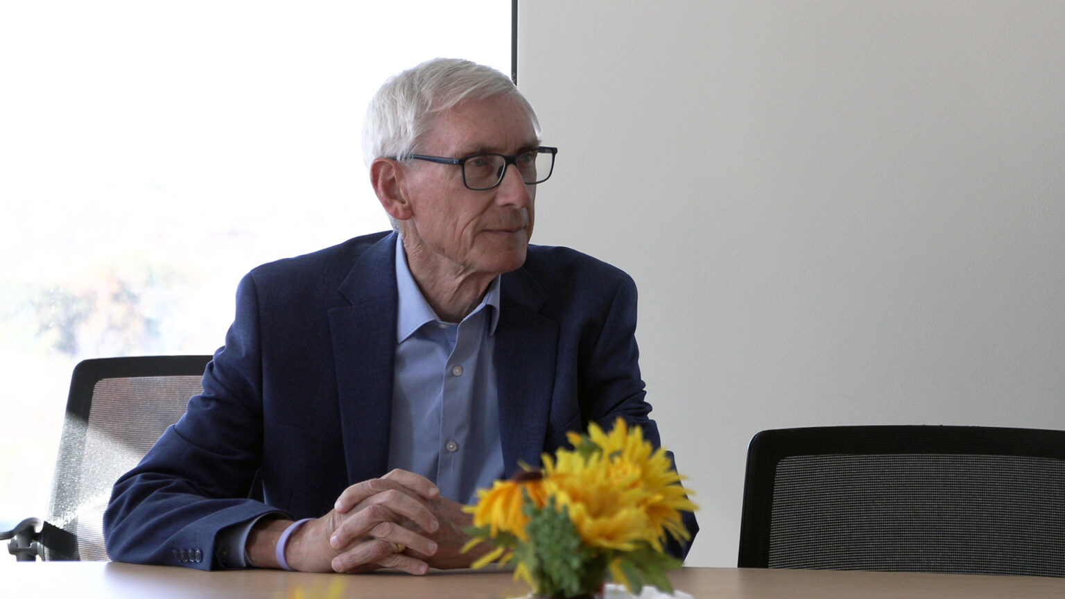 Tony Evers sits at a table with his hands folded on its surface, with an empty chair to one side, out-of-focus flowers in the foreground and sunlight illuminating a window in the background.