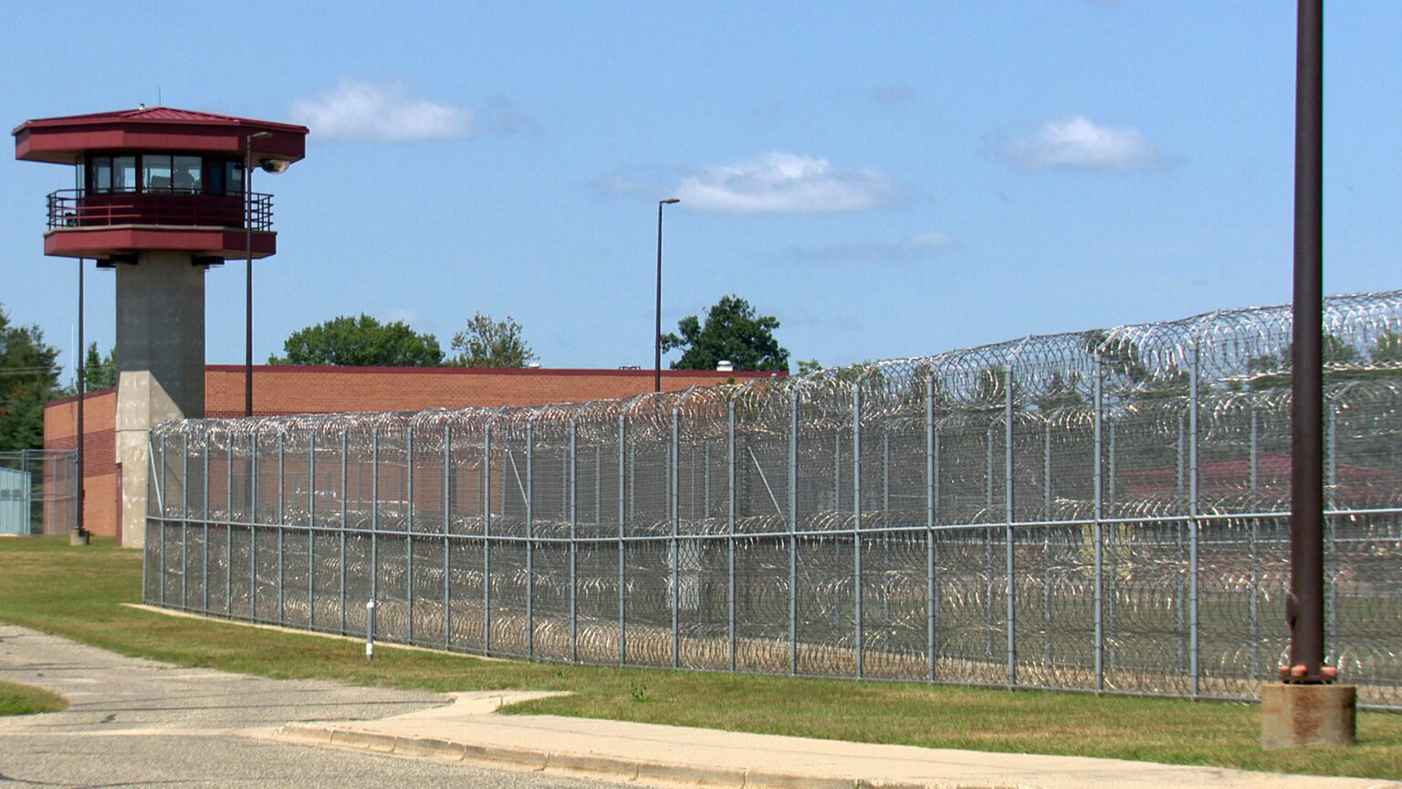 A chain-link fence ringed and topped with razor wire runs along the side of a parking lot, with a light pole in the foreground, and a prison guard tower, brick building, more lights and trees in the background, under a partly cloudy sky.