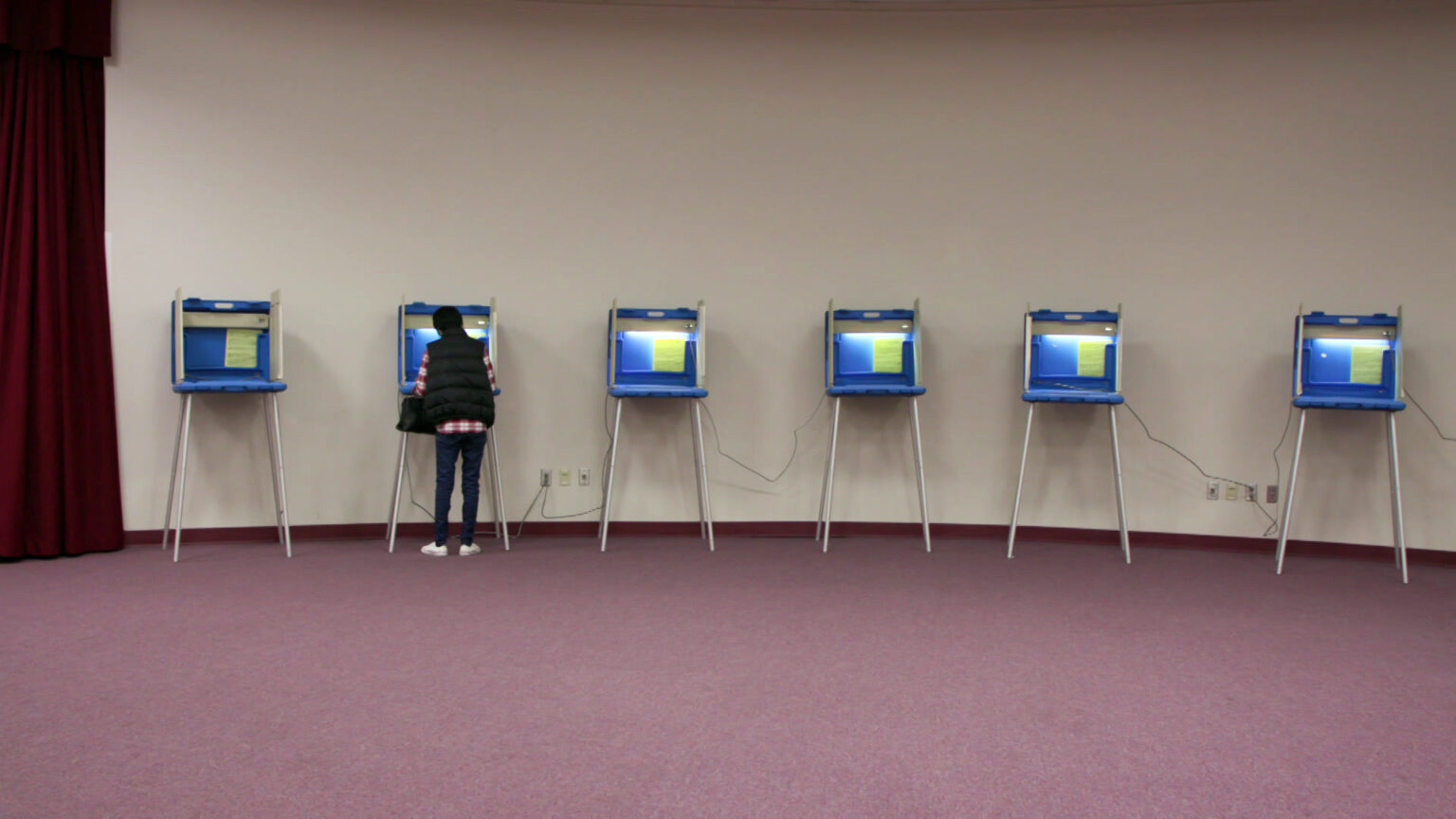 A voter fills out a ballot while standing and facing a voting booth with plastic privacy walls and metal legs, arranged in a row of similar booths in a room with a stage curtain on one side and a blank wall in the background.