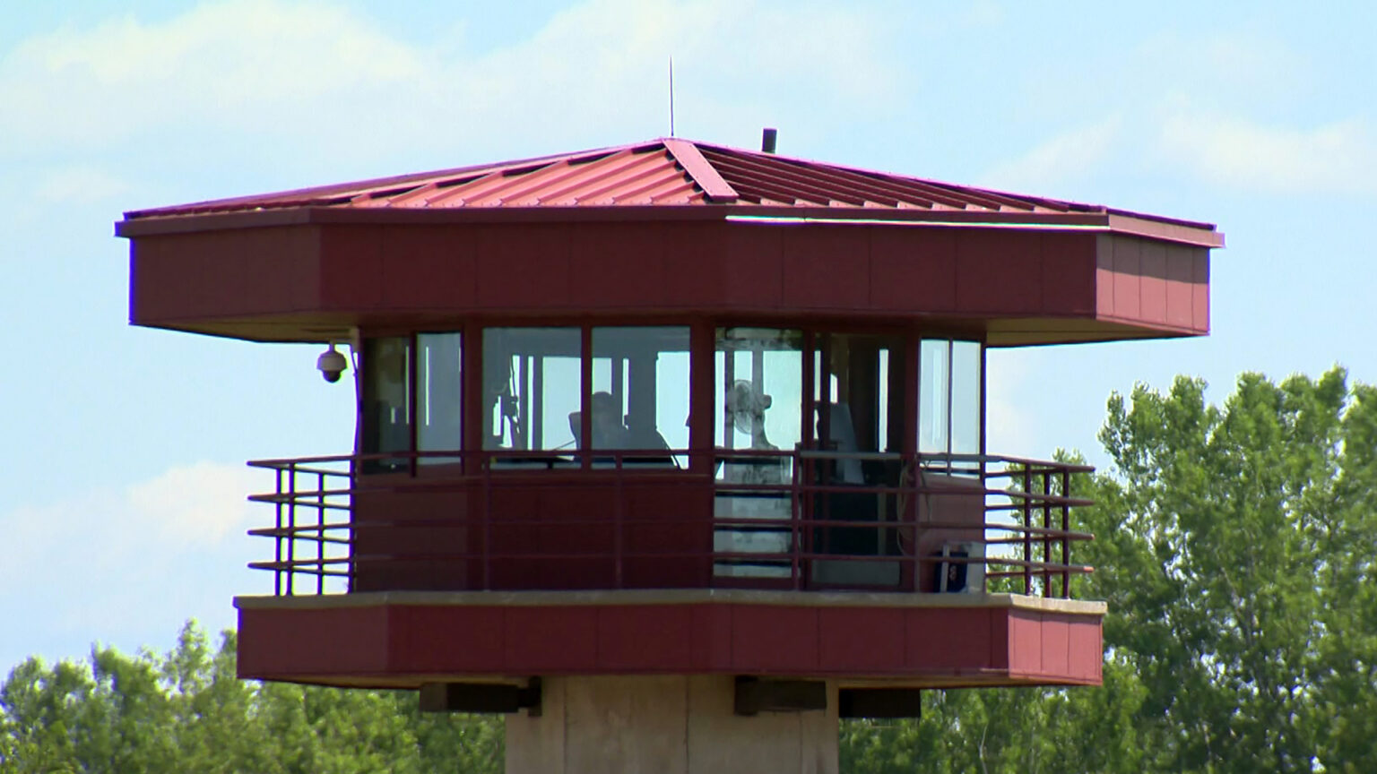 Corrections officers are visible through the windows of a prison guard tower with a security camera affixed to the underside of its low-slopped roof, with trees in the background.