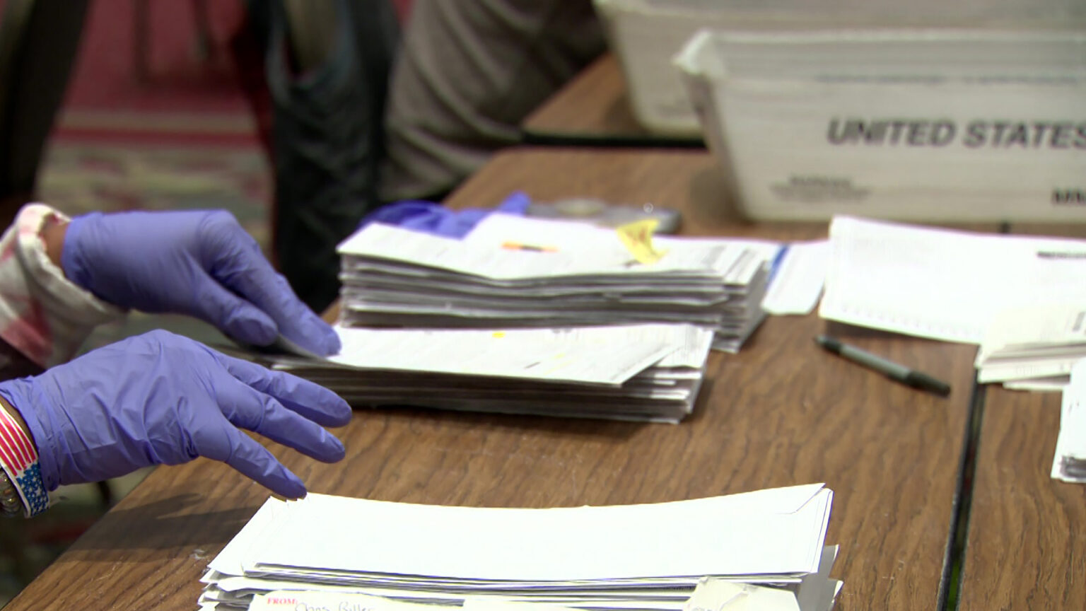 A pair of hands wearing nitrile gloves reaches toward a stack of envelopes on a laminated wood tabletop, with other stacks in the background alongside empty plastic U.S. mail trays.