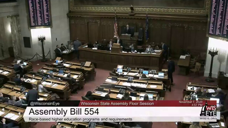 A video still image the floor of the Wisconsin Assembly with a legislative dais, rows of desks, two digital vote registers, the U.S. and Wisconsin flags, and a taxidermy bald eagle in a room with marble masonry, with a video graphic at bottom including the text Assembly Bill 554 and Race-based higher education programs and requirements.