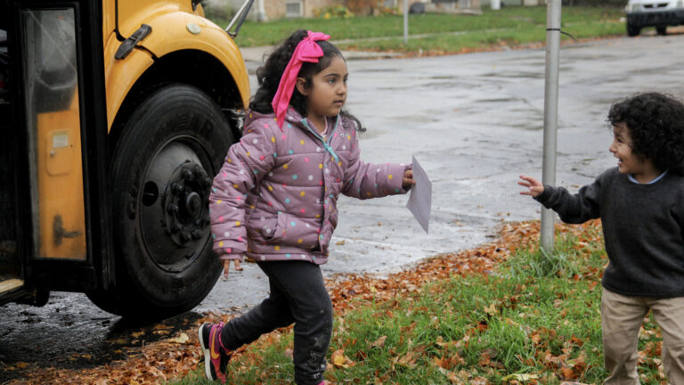 Magaly Coronado steps down from a school bus door on to leaf-covered turf and approaches Leonardo Coronado, who is reaching out to her with his right hand, with a rain-slicked street, a parked car and lawns in front of buildings in the background.