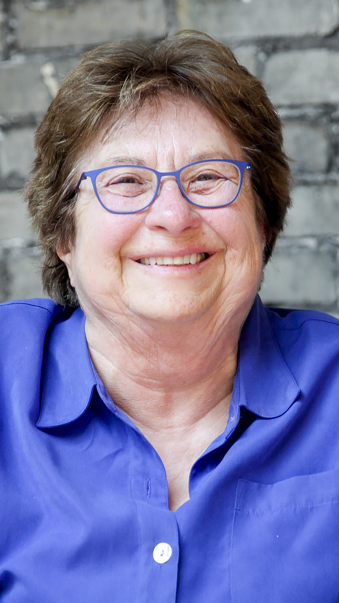 Sue Eckhart poses for a portrait while sitting in a room with a brick wall in the background.
