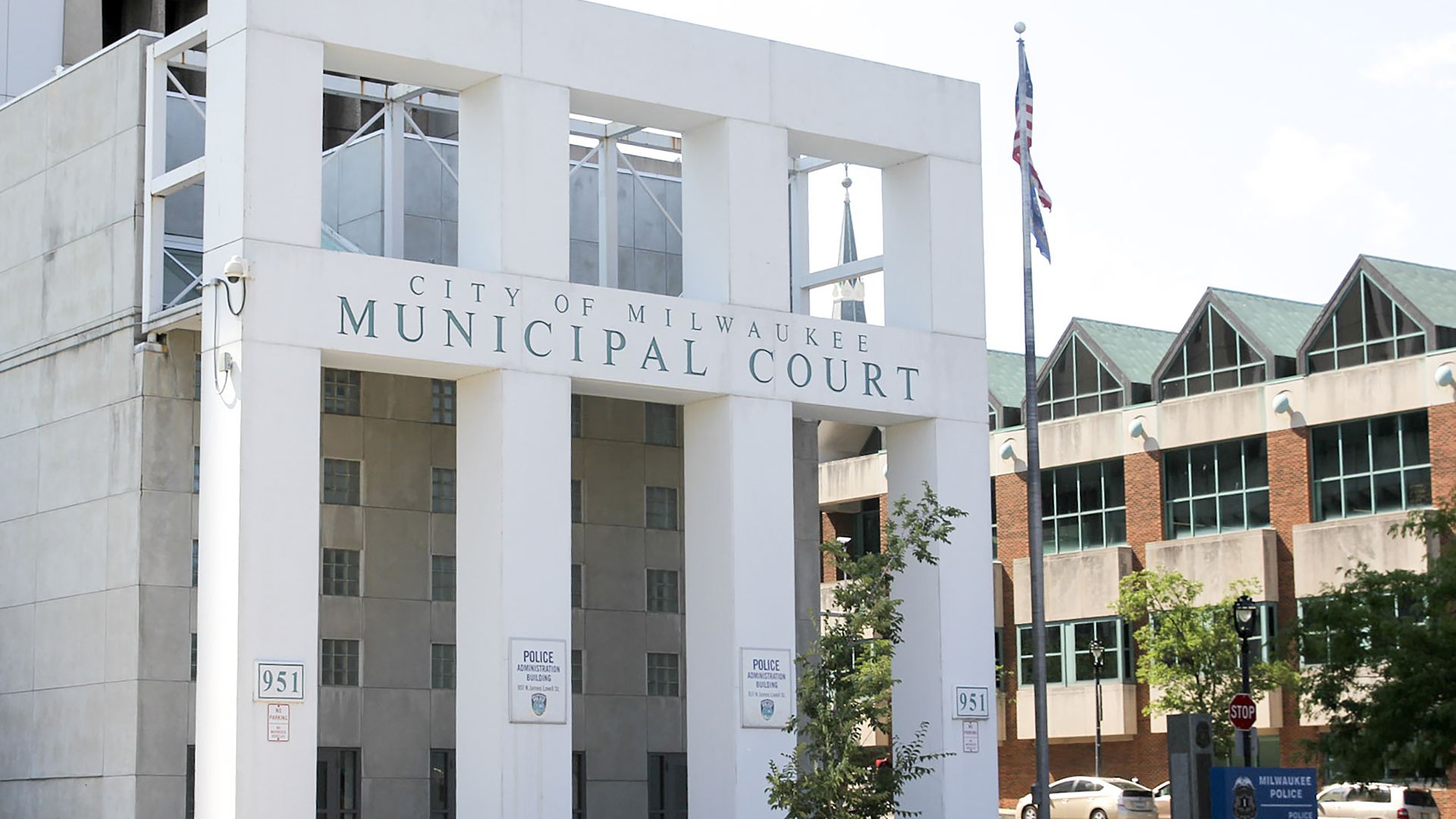 A concrete building with small windows is fronted by a facade with square pillars, a letter sign reading "City of Milwaukee Municipal Court," and smaller signs designating parking spots, with a flagpole, parked cars and another building in the background.