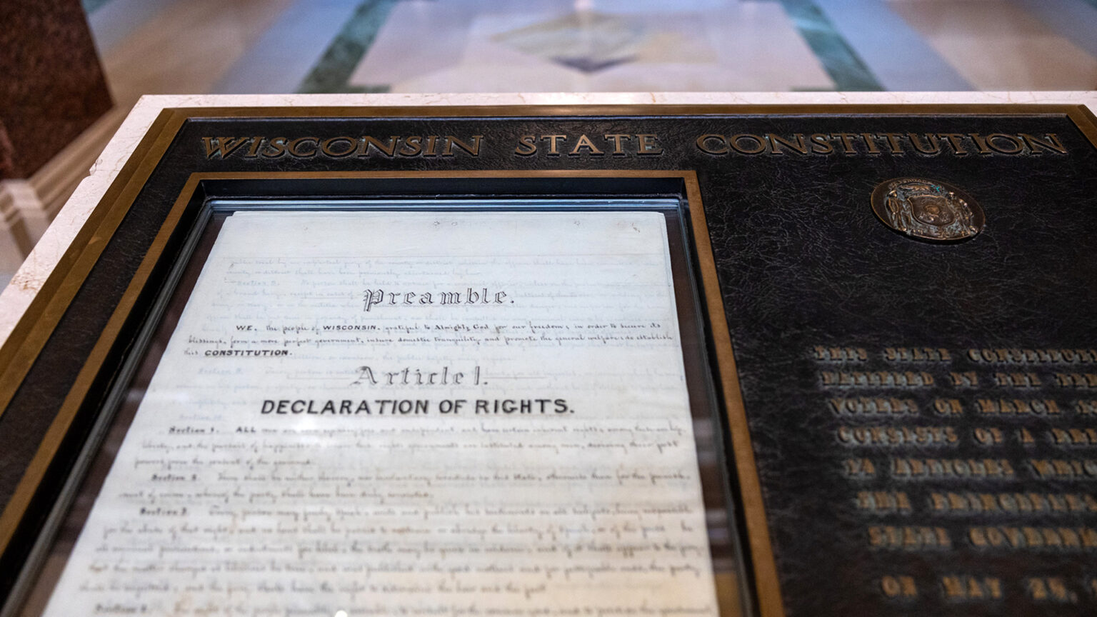 A historical display shows a bronze plaque with the title Wisconsin State Constitution and a glass-covered set of papers with header copy on the top reading Preamble and Article I. Declaration of Rights, with an out-of-focus marble floor in the background.