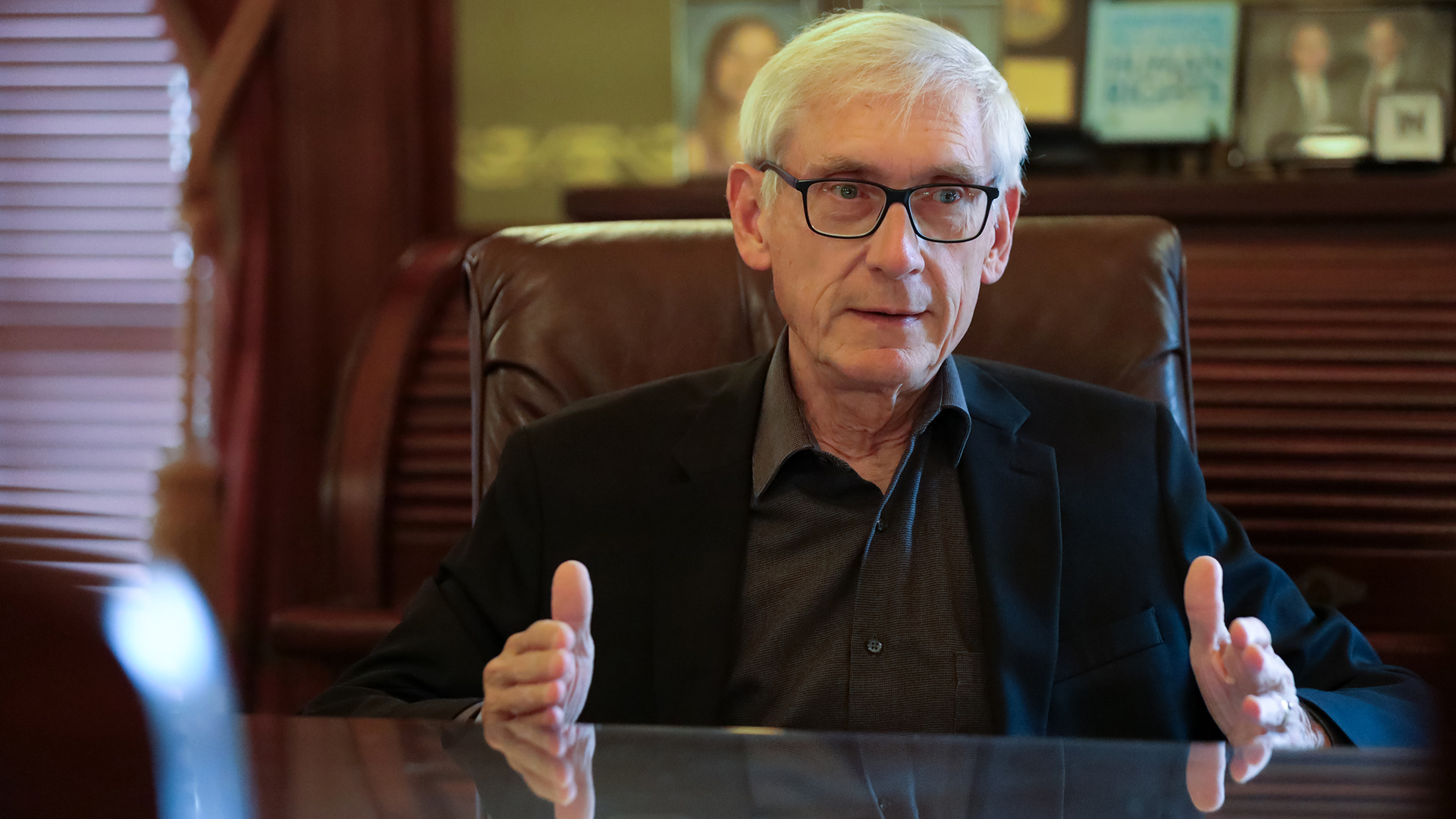 Tony Evers sits in a high-backed leather chair and gestures with both hands on a glass-topped table, with a roll top desk with photos and other items on it in the background.