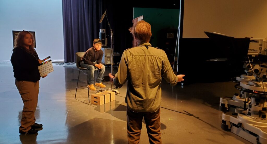 A media production professional speaks with two high school students who are about to conduct an interview in a studio.