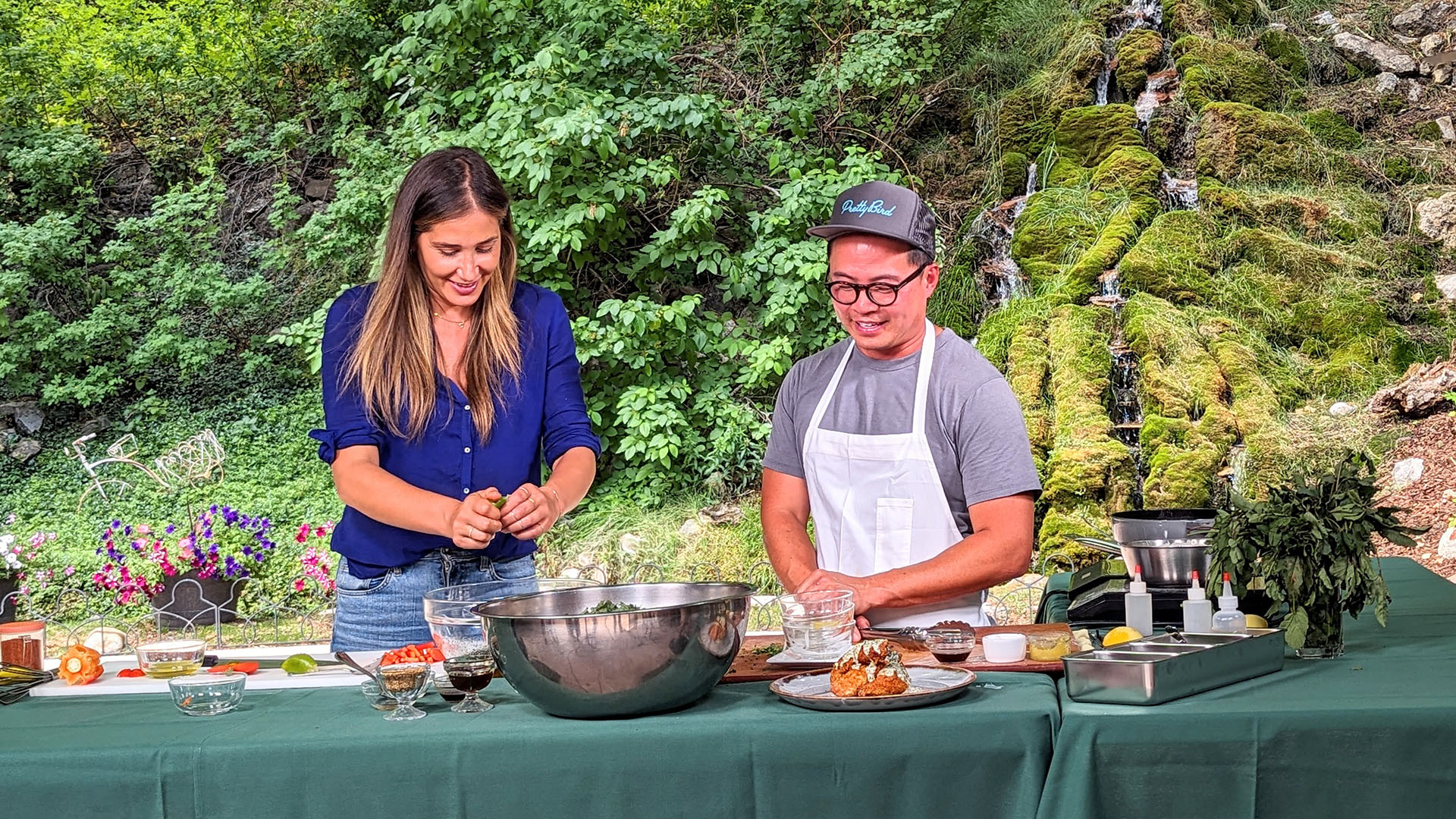 A man and woman combine ingredients in bowls on a table while doing a cooking demonstration outdoors.