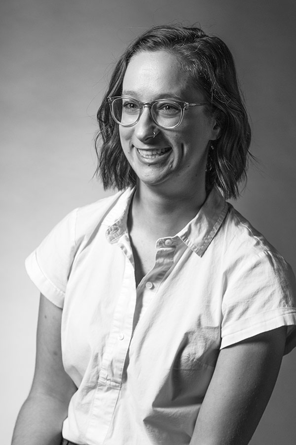 Black and white photo of a smiling woman with glasses, a nose ring, and short hair, wearing a white shirt.