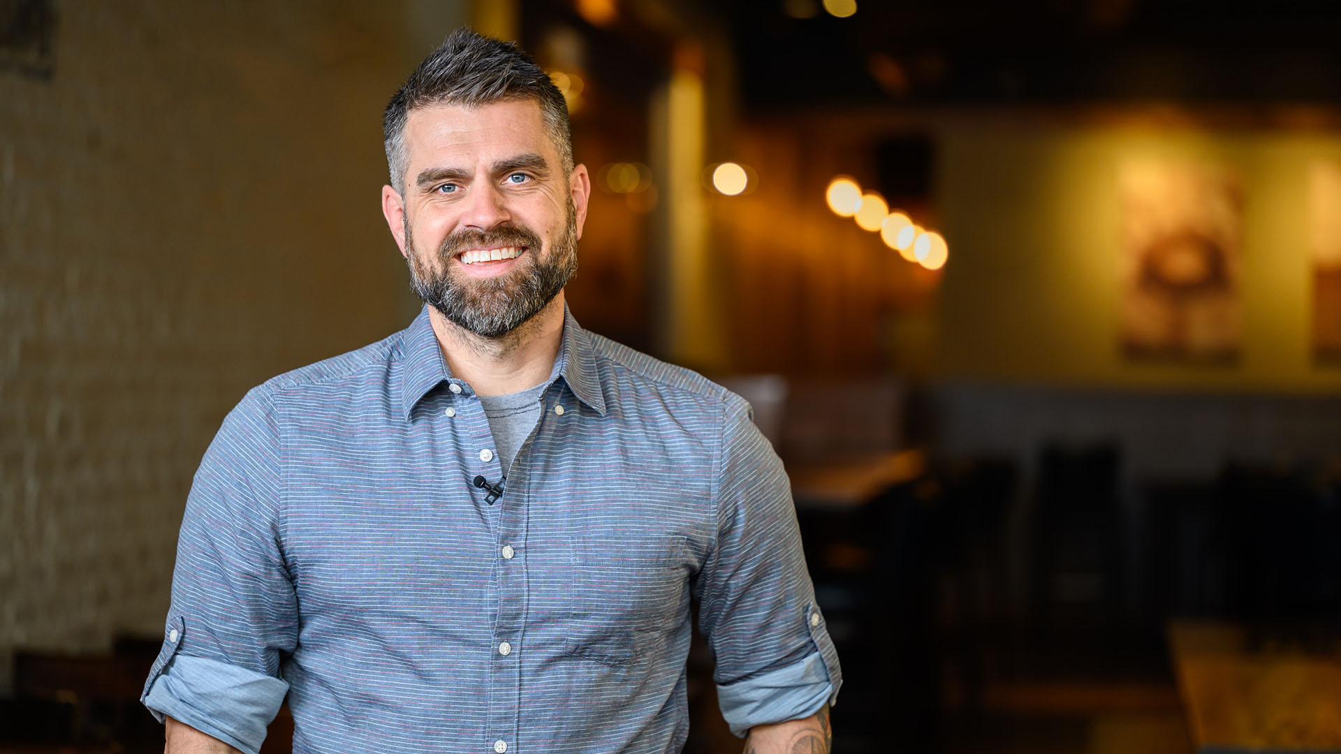 Wisconsin Foodie host Luke Zahm smiles at the camera from within a low-lit restaurant and bar.
