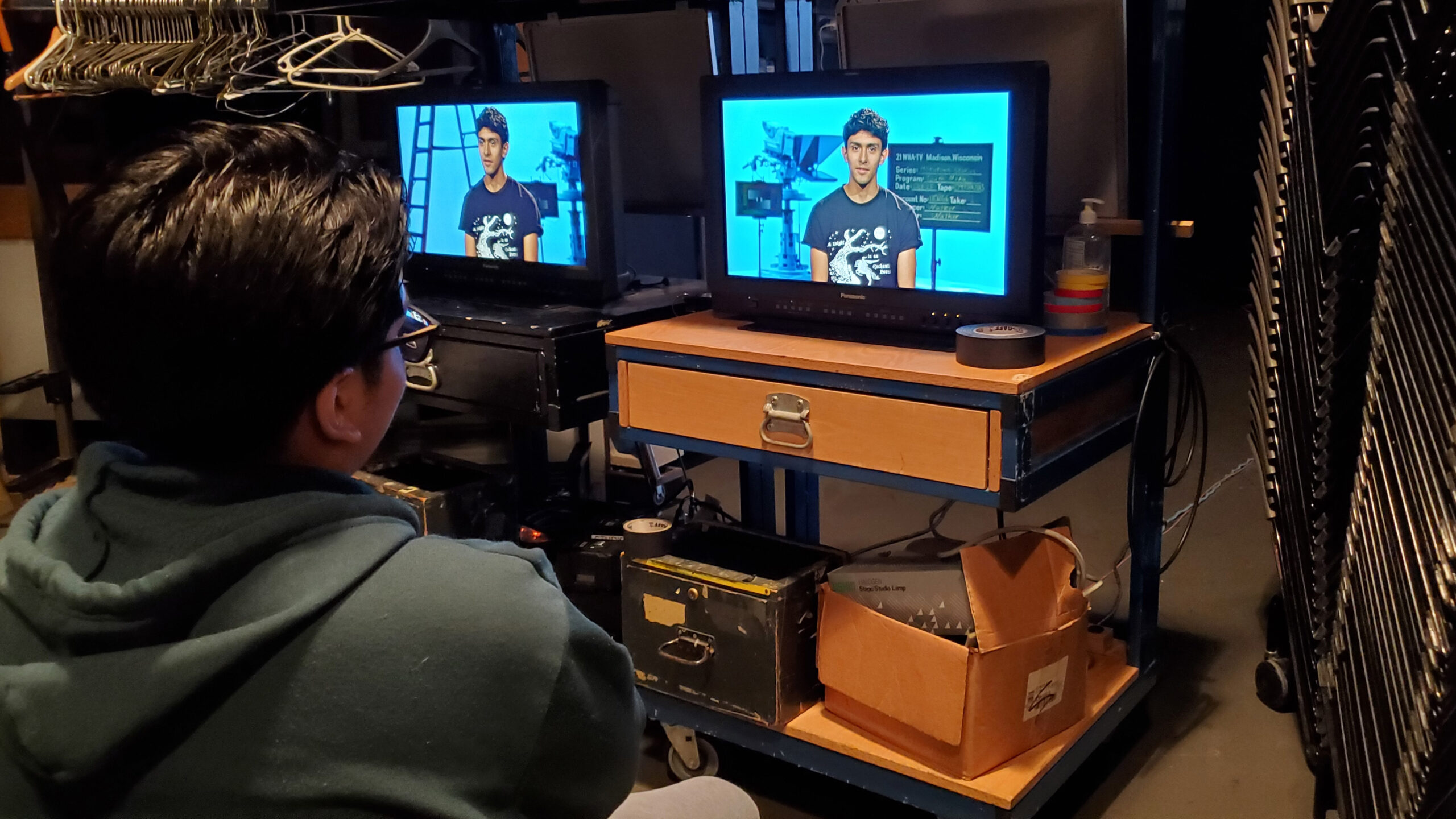 A high school student watches monitors showing another student in a production studio.