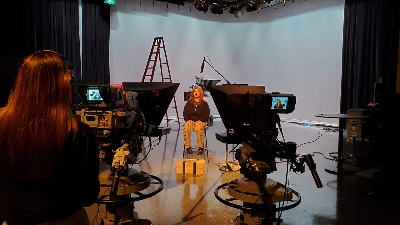 A person sitting on a stool in a studio setup with professional video cameras pointed towards them.