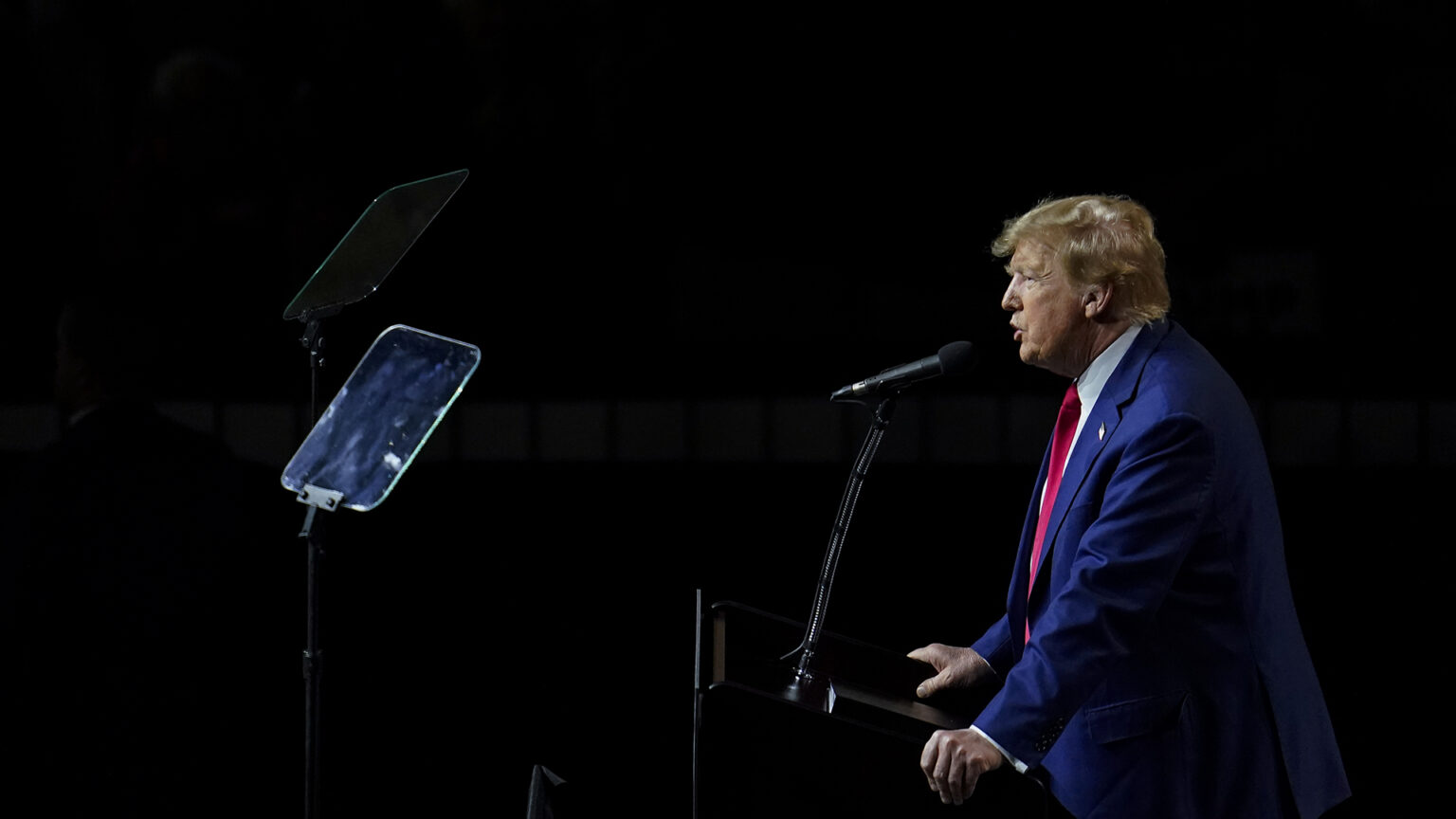 Donald Trump speaks into a microphone mounted to a podium which he is holding with both hands while reading from two teleprompters.