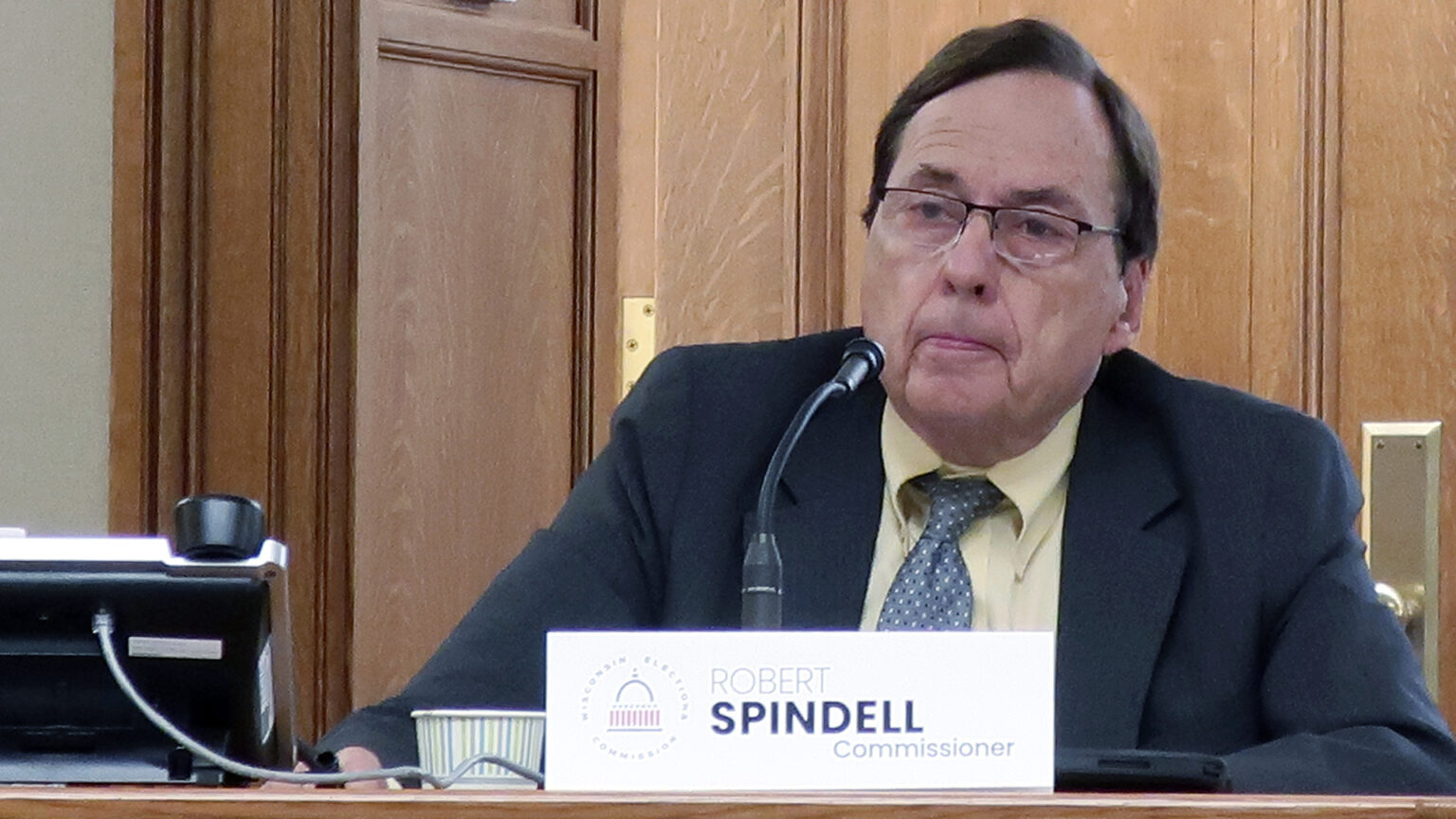 Robert Spindell sits and speaks into a microphone on a table with a corded office phone, paper cup and name sign reading Robert Spindell and Commissioner on its surface, with wood doors in the background.