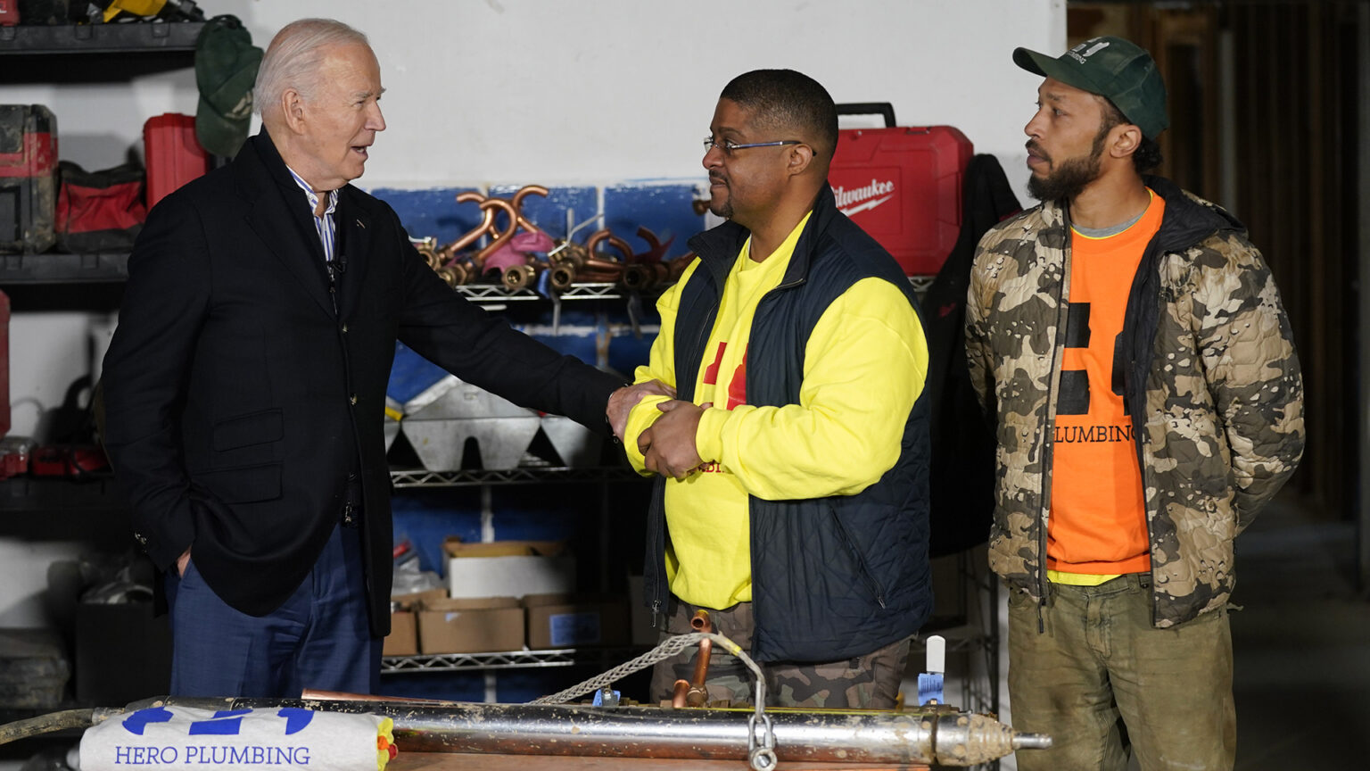 Joe Biden holds the wrist of a man facing him with his hands clasped, and with another man to the side standing with arms behind his back, in a room with plumbing equipment on a table and on shelves along the rear wall.